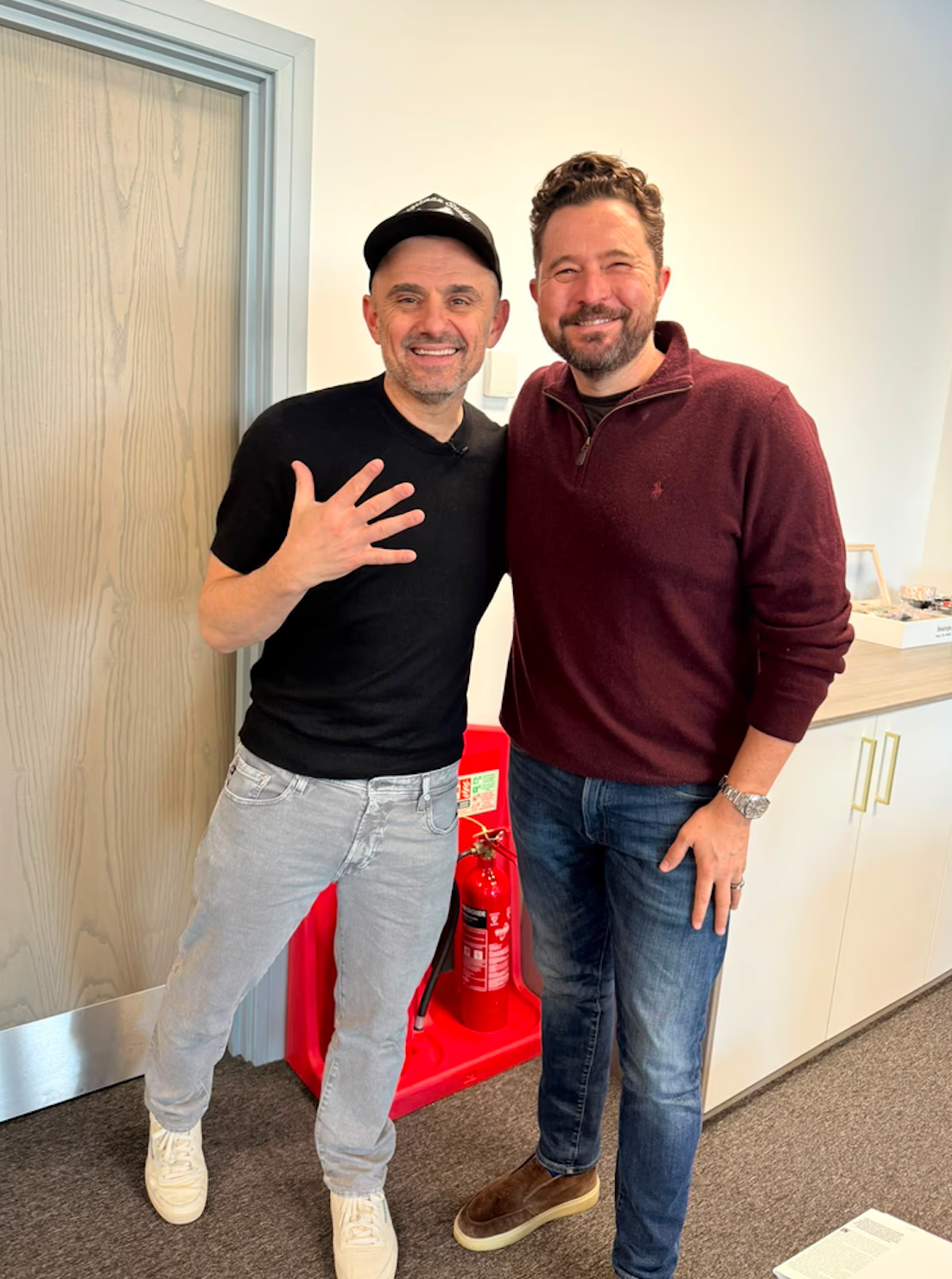 Two men smiling together in an office setting, one wearing a black shirt and the other in a maroon sweater.