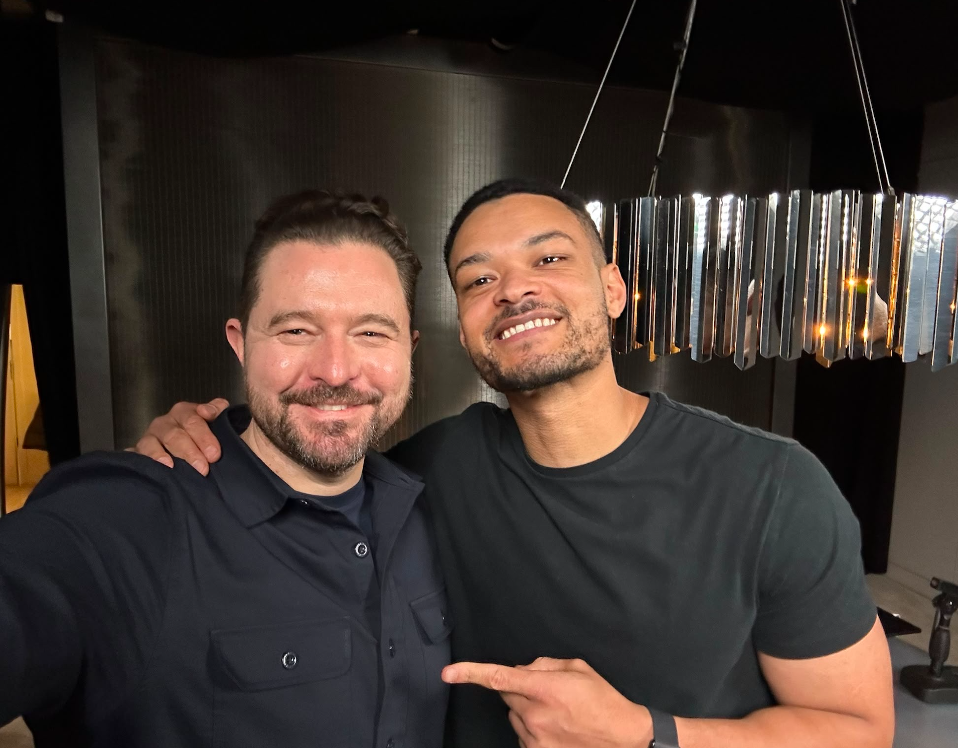 Two men smiling and posing together in a modern indoor setting with stylish lighting.