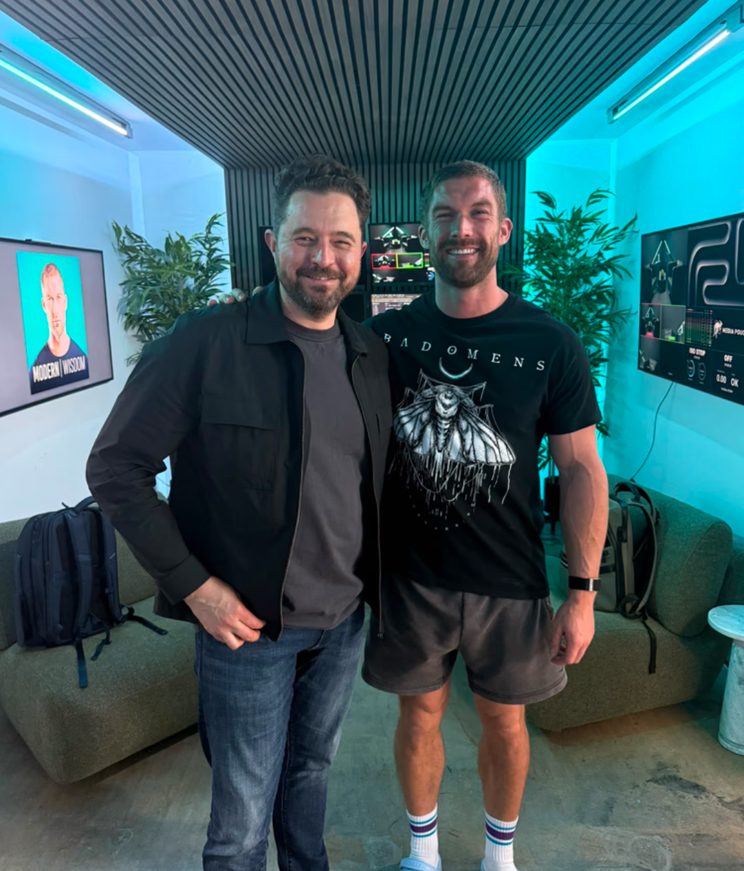 Two men pose together in a modern indoor setting with green plants and digital screens in the background.