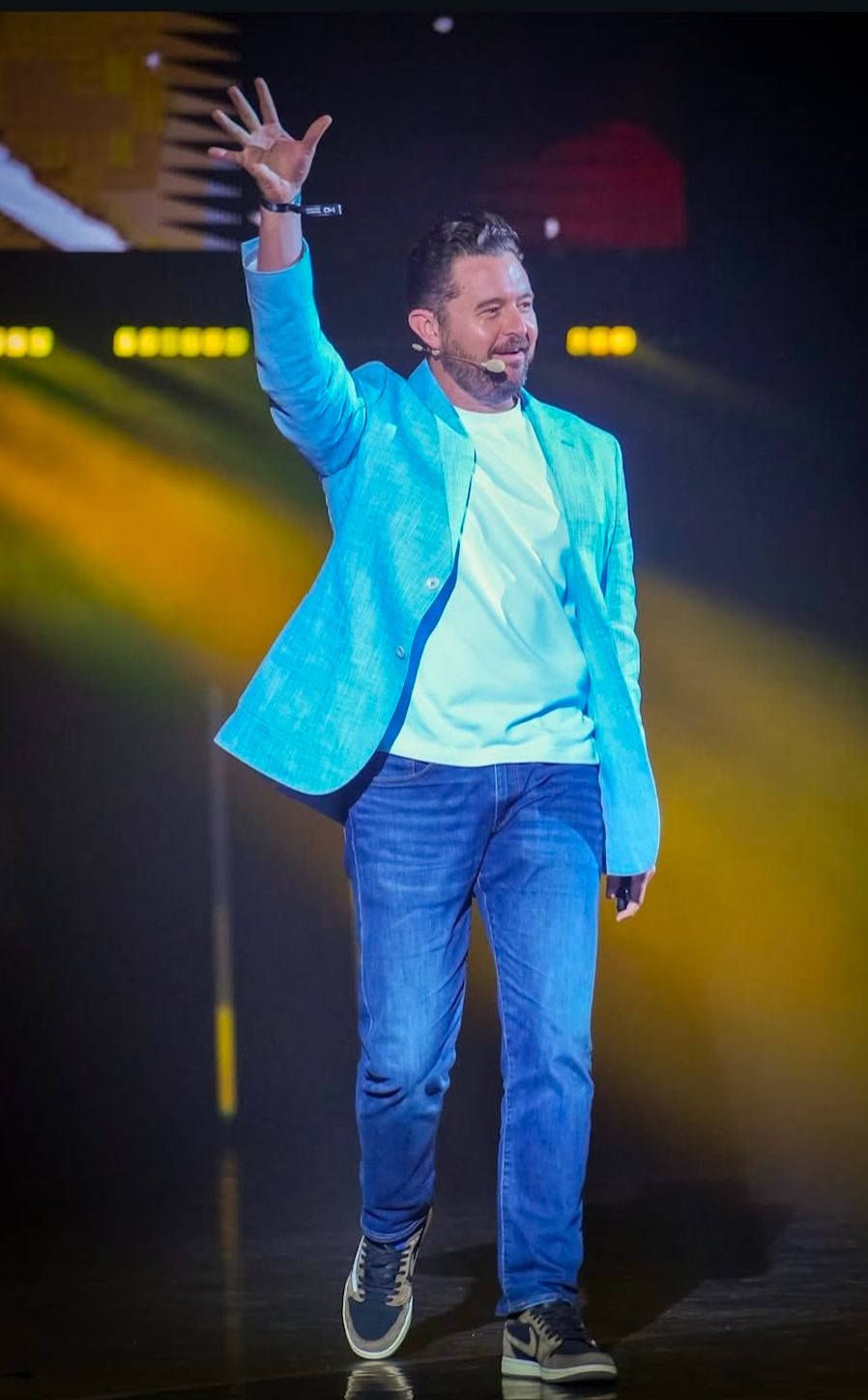A man in a blue blazer and jeans gestures enthusiastically while on stage, illuminated by bright lights.