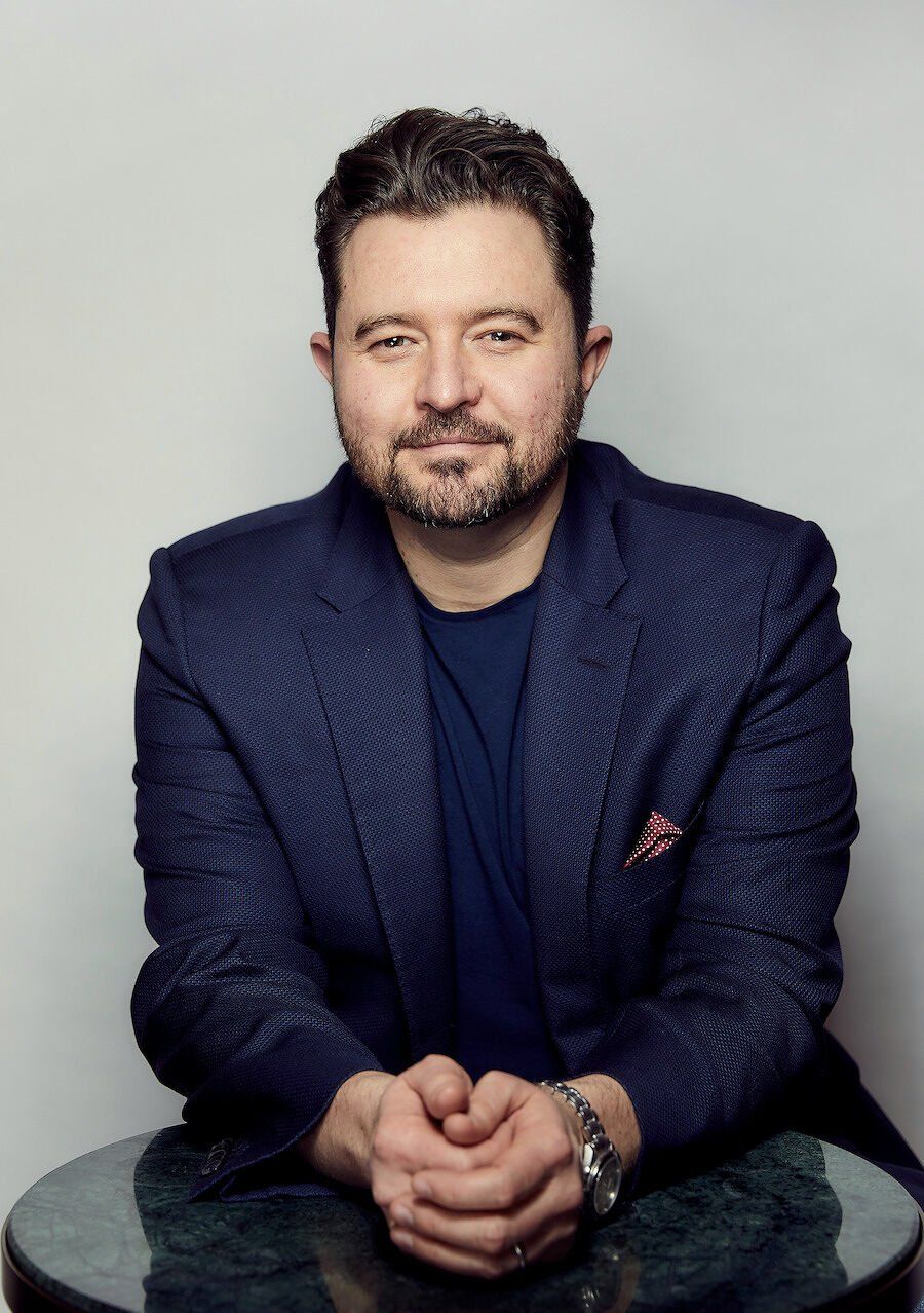 A man with a beard and short hair is seated at a table, wearing a navy blazer over a dark shirt, with a confident expression.