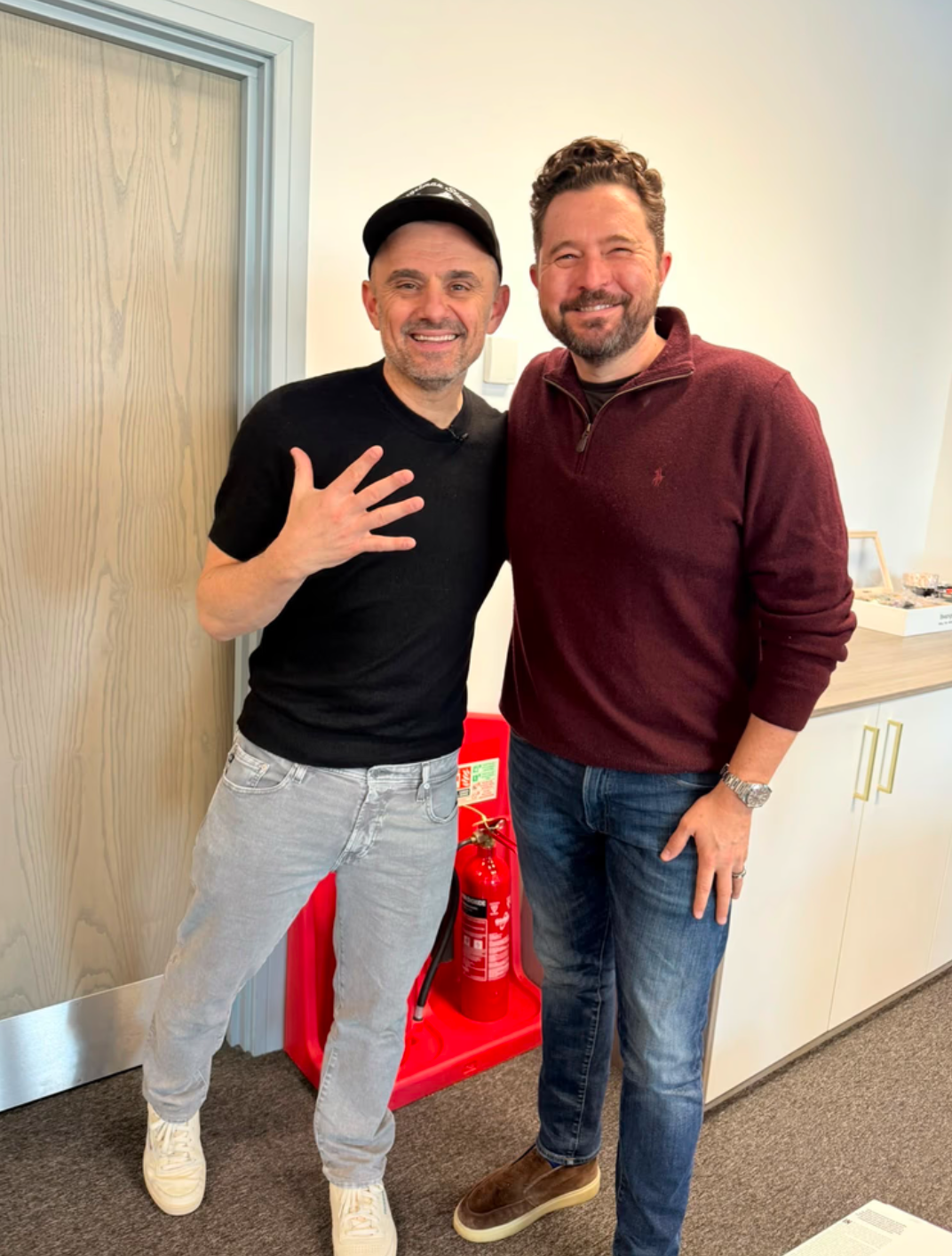 Two men smiling and posing together in an office setting, one wearing a black shirt and the other in a maroon sweater.