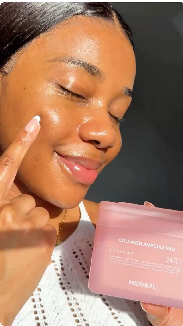 A woman with glowing skin holds a pink skincare product while pointing to her cheek, showcasing her healthy complexion.