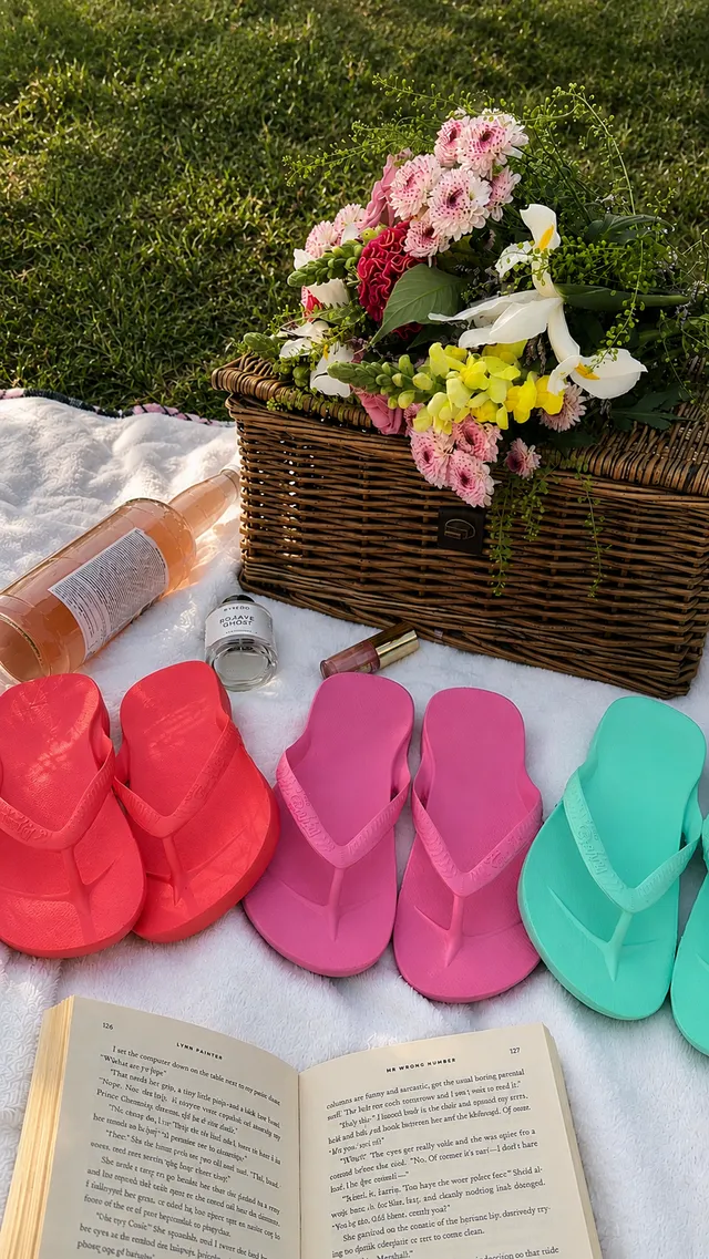 A vibrant picnic setup featuring colorful flip-flops arranged in a row, a wicker basket filled with flowers, a bottle of rosé, a perfume bottle, and an open book on a white blanket.