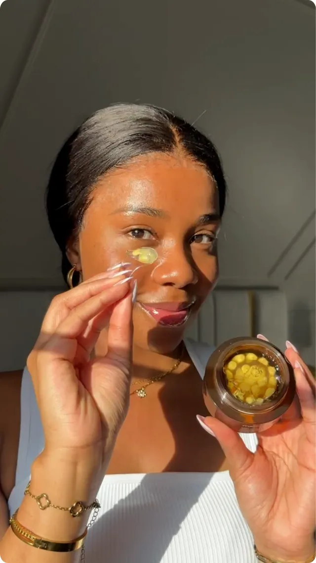A woman holds a skincare product close to her face, applying a gel-like substance. She has a radiant smile and is illuminated by natural light.