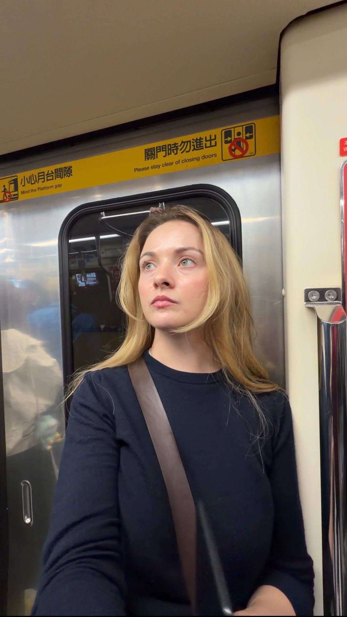 A woman with long blonde hair stands inside a subway train, gazing thoughtfully out of the window. The interior features metallic surfaces and signage in multiple languages.