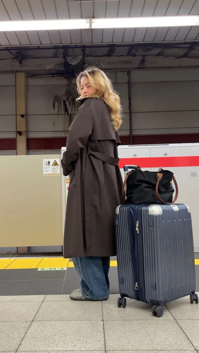 A woman stands at a train station, wearing a brown coat and blue jeans, with a suitcase beside her and a black bag in hand.