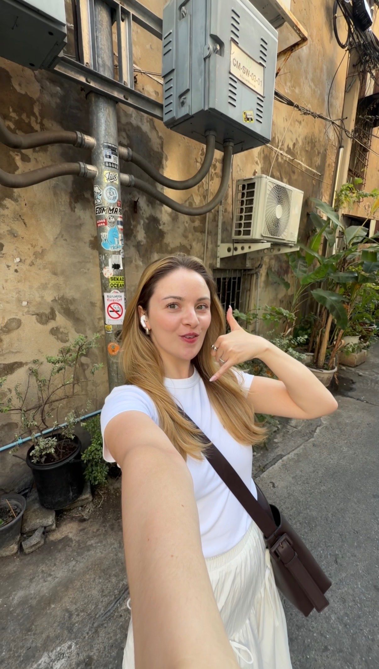 A woman taking a selfie on a street with a mix of urban elements, including air conditioning units and greenery.