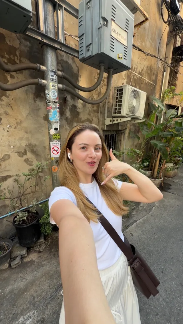 A woman taking a selfie on a street with a mix of urban elements, including air conditioning units and greenery.