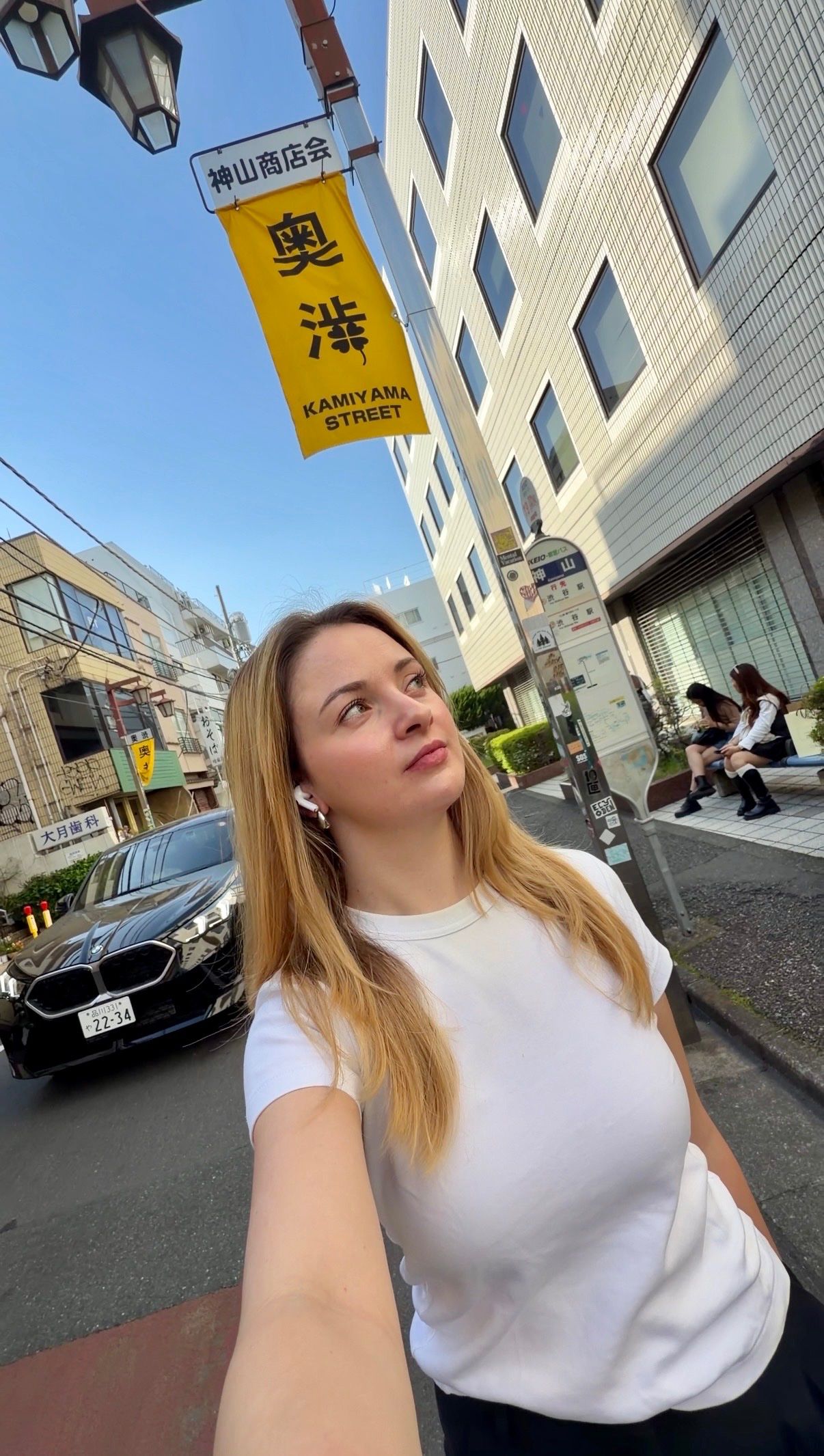A woman stands on a street in front of a sign that reads 'Kamiyama Street', with buildings and people in the background.