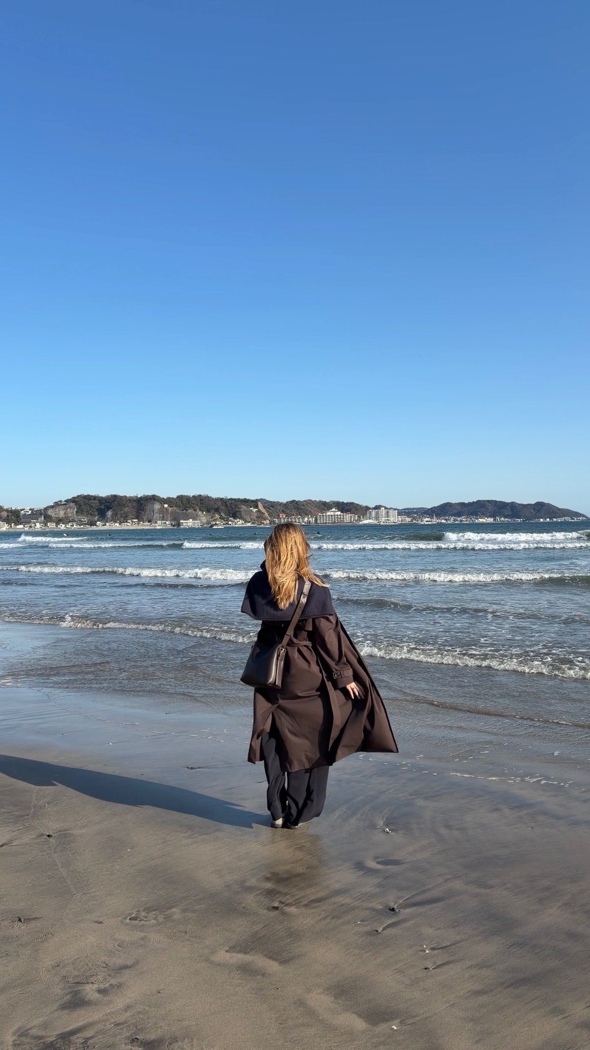 A person with long hair walks along the beach, facing the ocean under a clear blue sky.