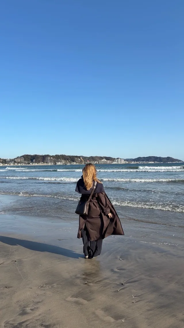 A person with long hair walks along the beach, facing the ocean under a clear blue sky.