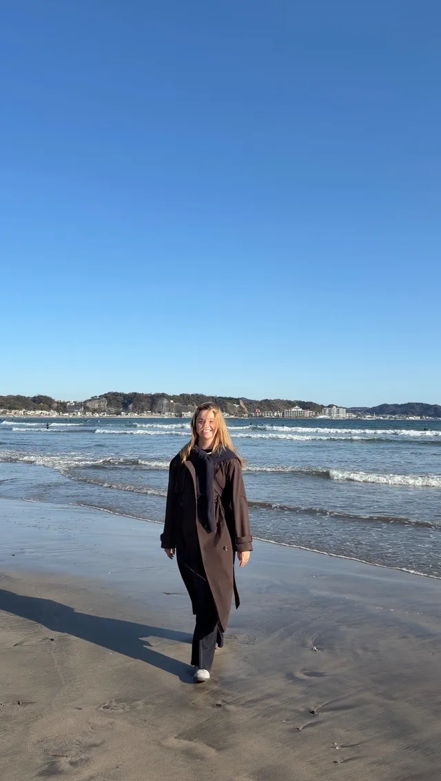 A woman walks along the beach, wearing a long coat and scarf, with waves and a clear blue sky in the background.