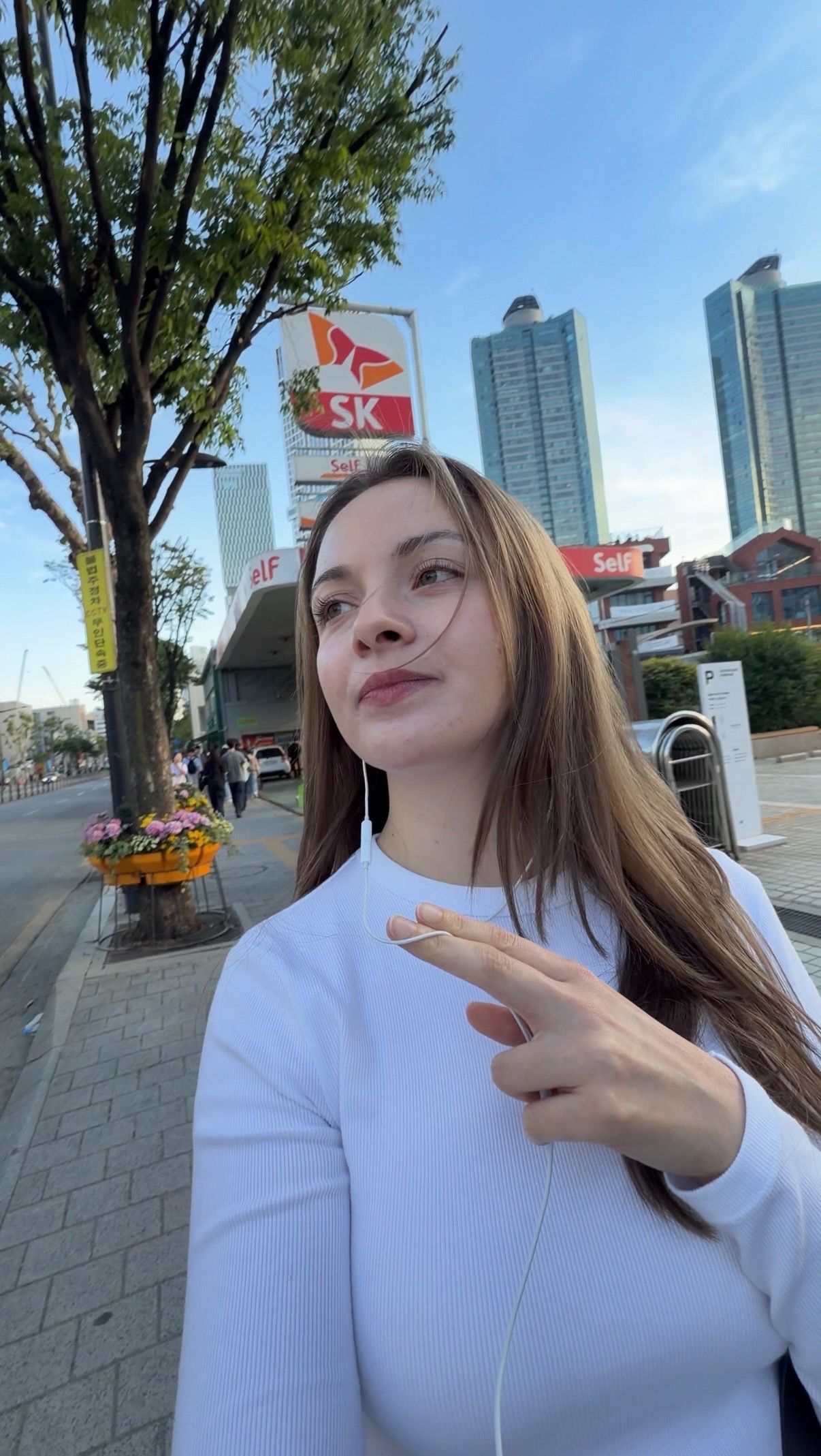A young woman poses on a city street, wearing a white long-sleeve top and earbuds, with a confident expression and a peace sign gesture.
