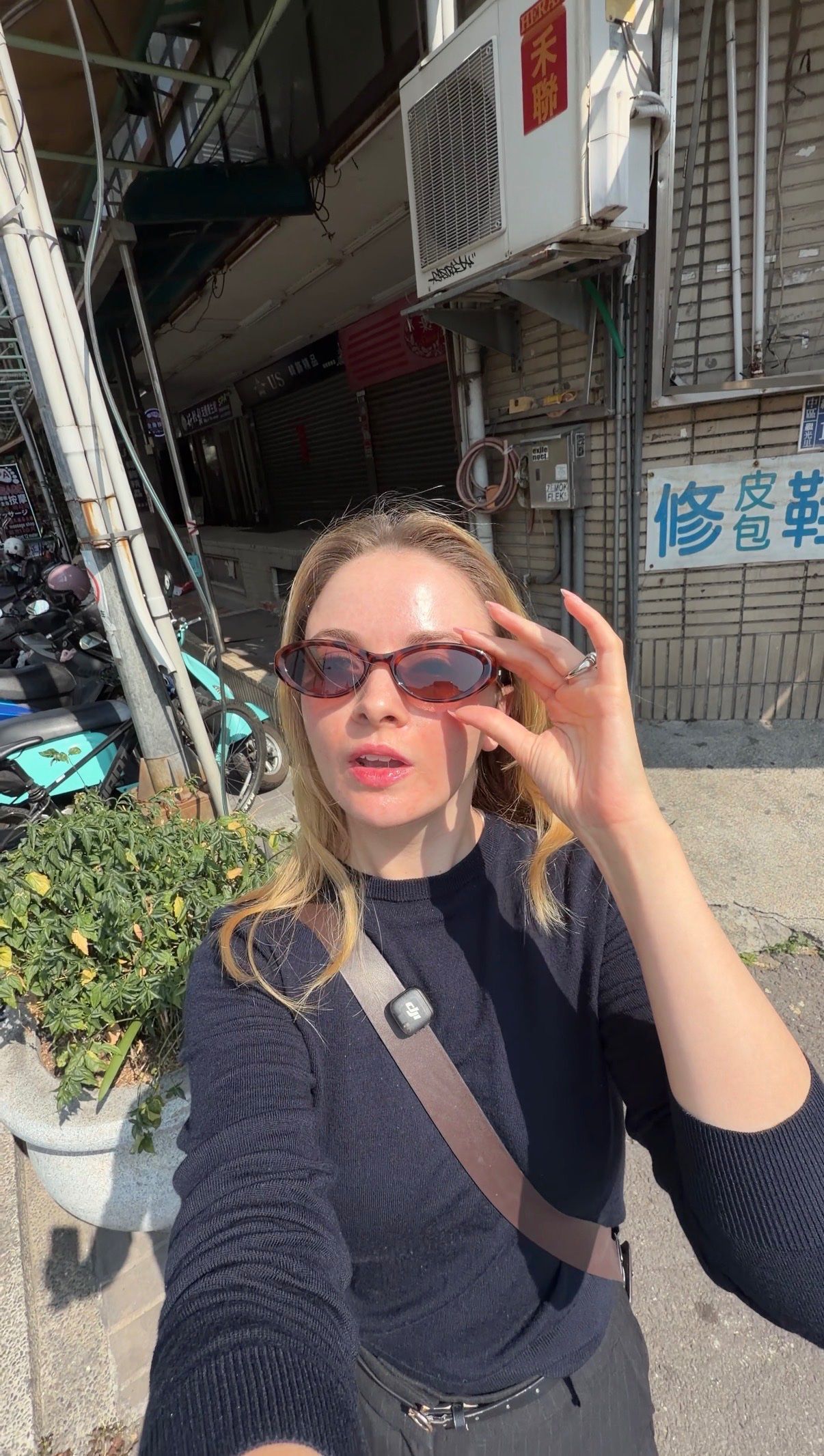 A person wearing stylish sunglasses poses outdoors, with a street scene and parked scooters in the background.