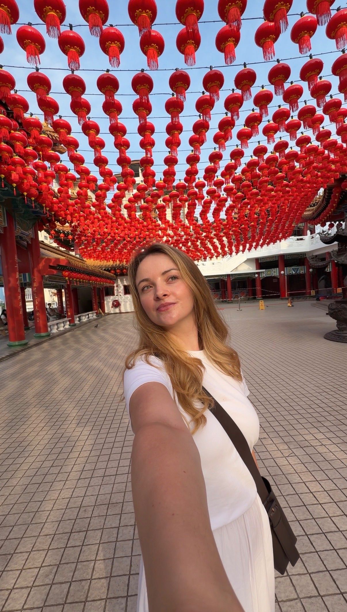 A woman poses for a selfie in a vibrant setting adorned with numerous red lanterns hanging overhead.