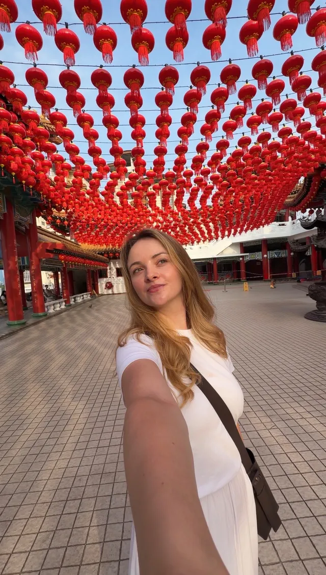 A woman poses for a selfie in a vibrant setting adorned with numerous red lanterns hanging overhead.