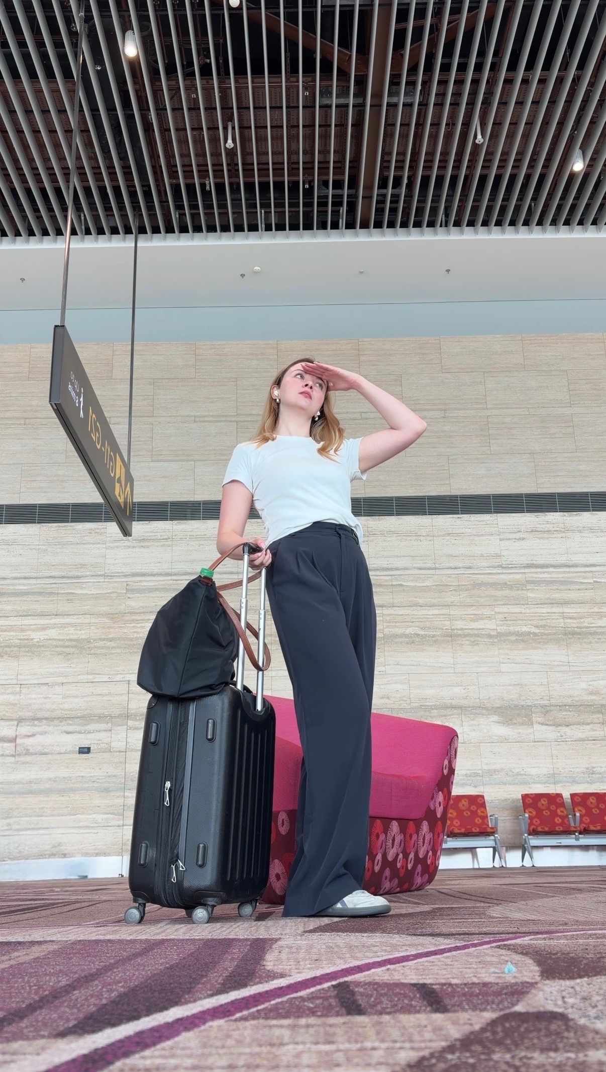A traveler stands in an airport terminal, looking out as they prepare to depart, with a suitcase beside them.