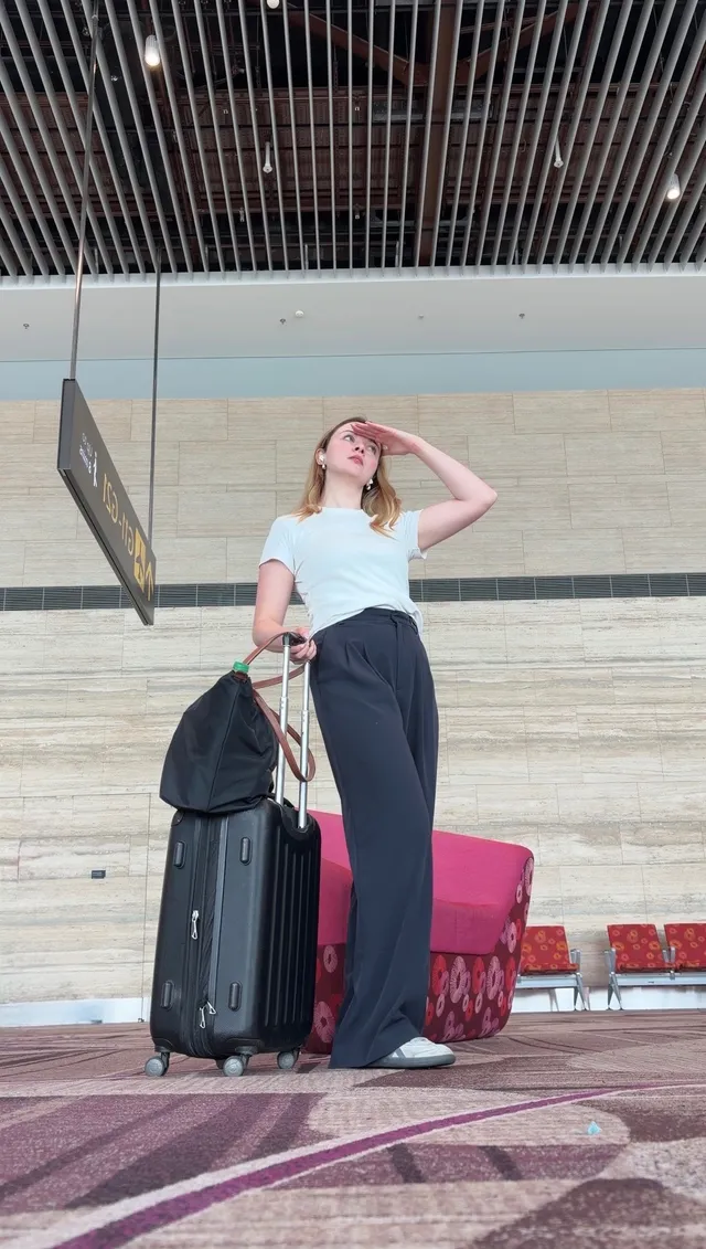 A traveler stands in an airport terminal, looking out as they prepare to depart, with a suitcase beside them.