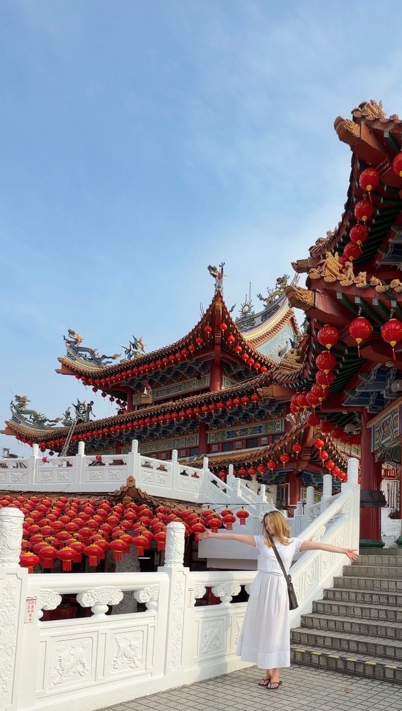 A woman in a white dress stands with her arms outstretched in front of a traditional Chinese temple adorned with red lanterns.