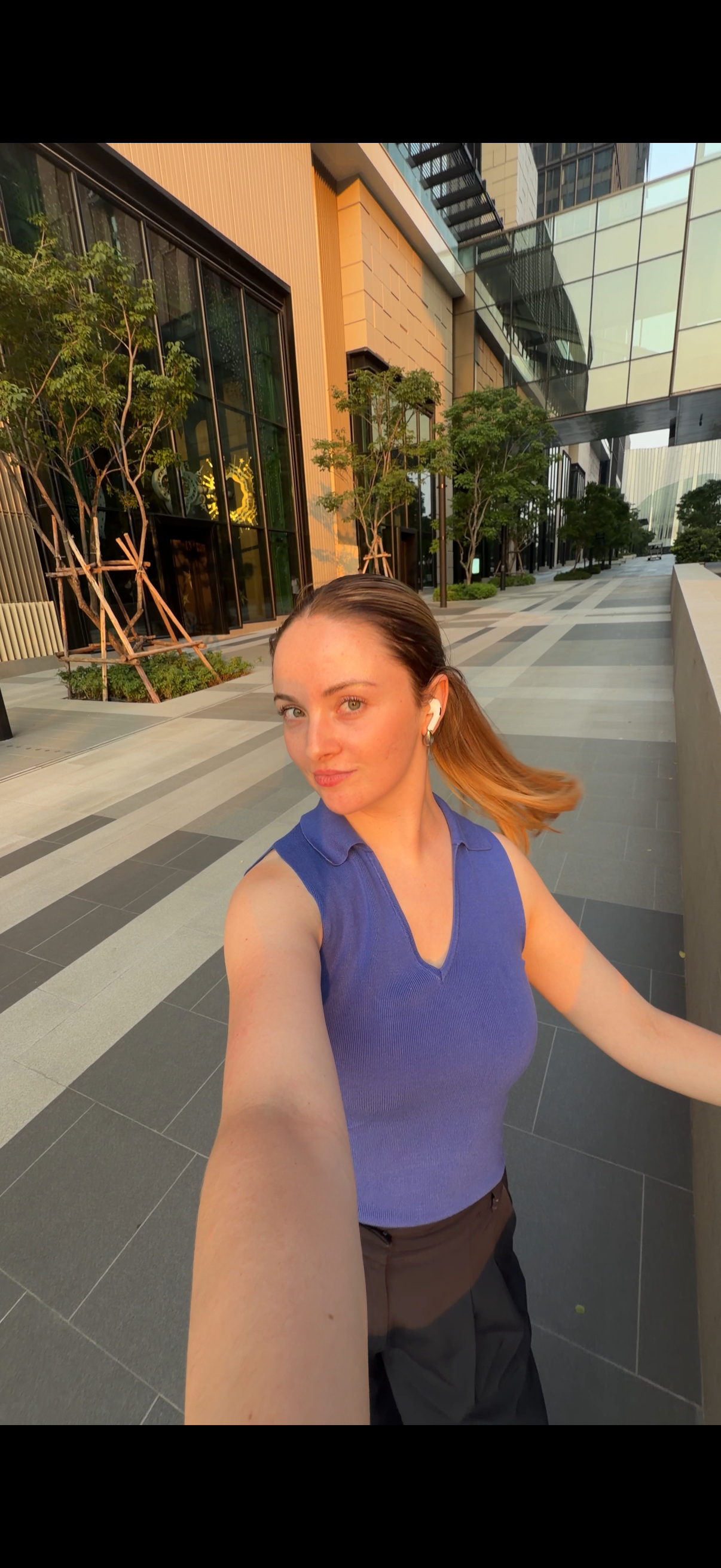 A young woman poses for a selfie in an urban setting, wearing a blue top and wireless earbuds, with modern architecture and greenery in the background.