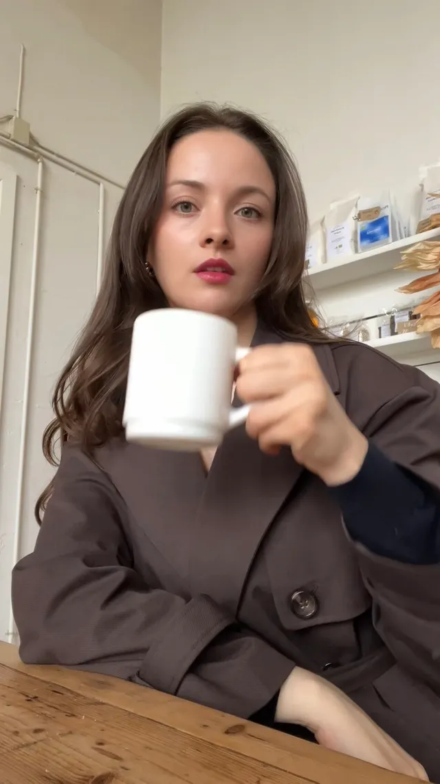 A woman with long hair holds a white mug while sitting at a wooden table, wearing a stylish brown coat.
